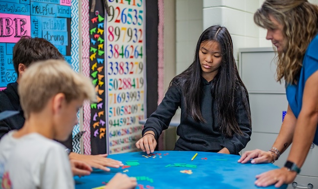 students working on a hands-on activity around a table while a teacher watches